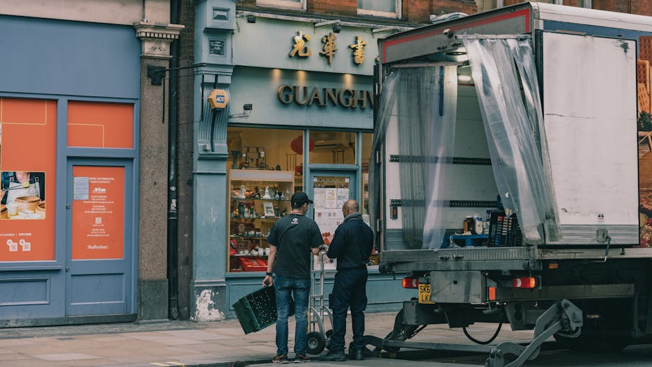 Two men dressed in dark clothing stand on a city sidewalk near a partially loaded moving truck with a large open rear door, revealing moving supplies and packing materials inside. They are next to a s
